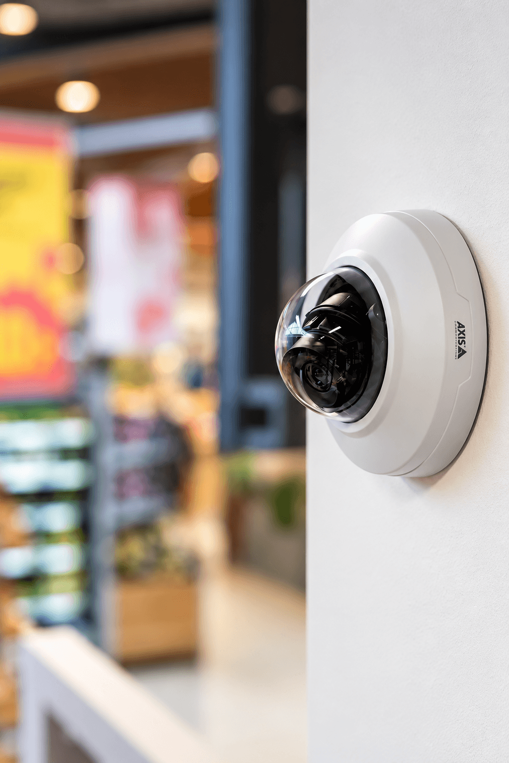 White Axis dome security camera mounted on a wall overlooking a blurred retail store interior.